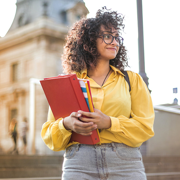 Female college student standing on campus carrying books and wearing a backpack.